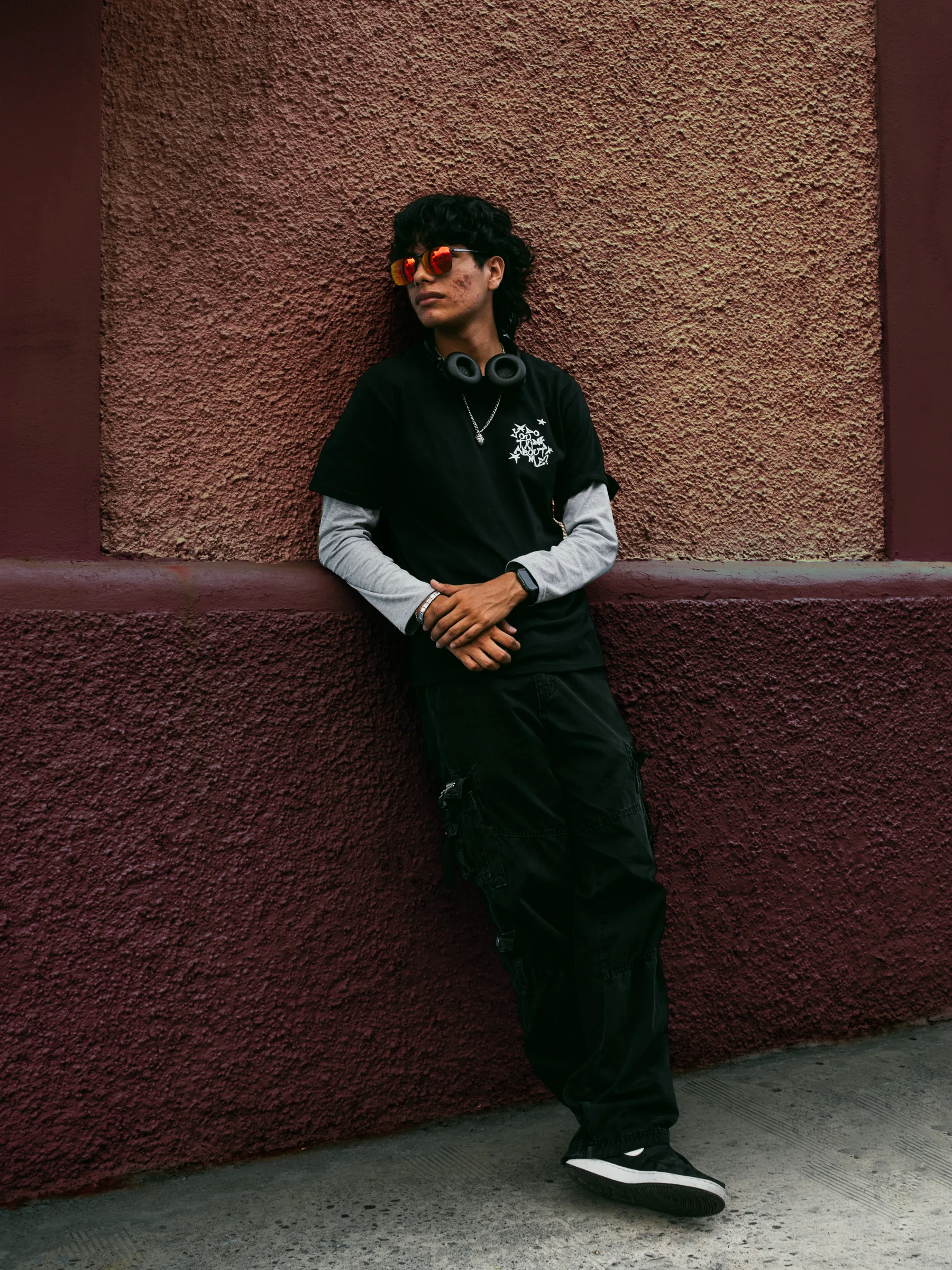 Modelo con lentes de sol y audífonos alrededor del cuello, recargado en una pared, con playera negra del merch