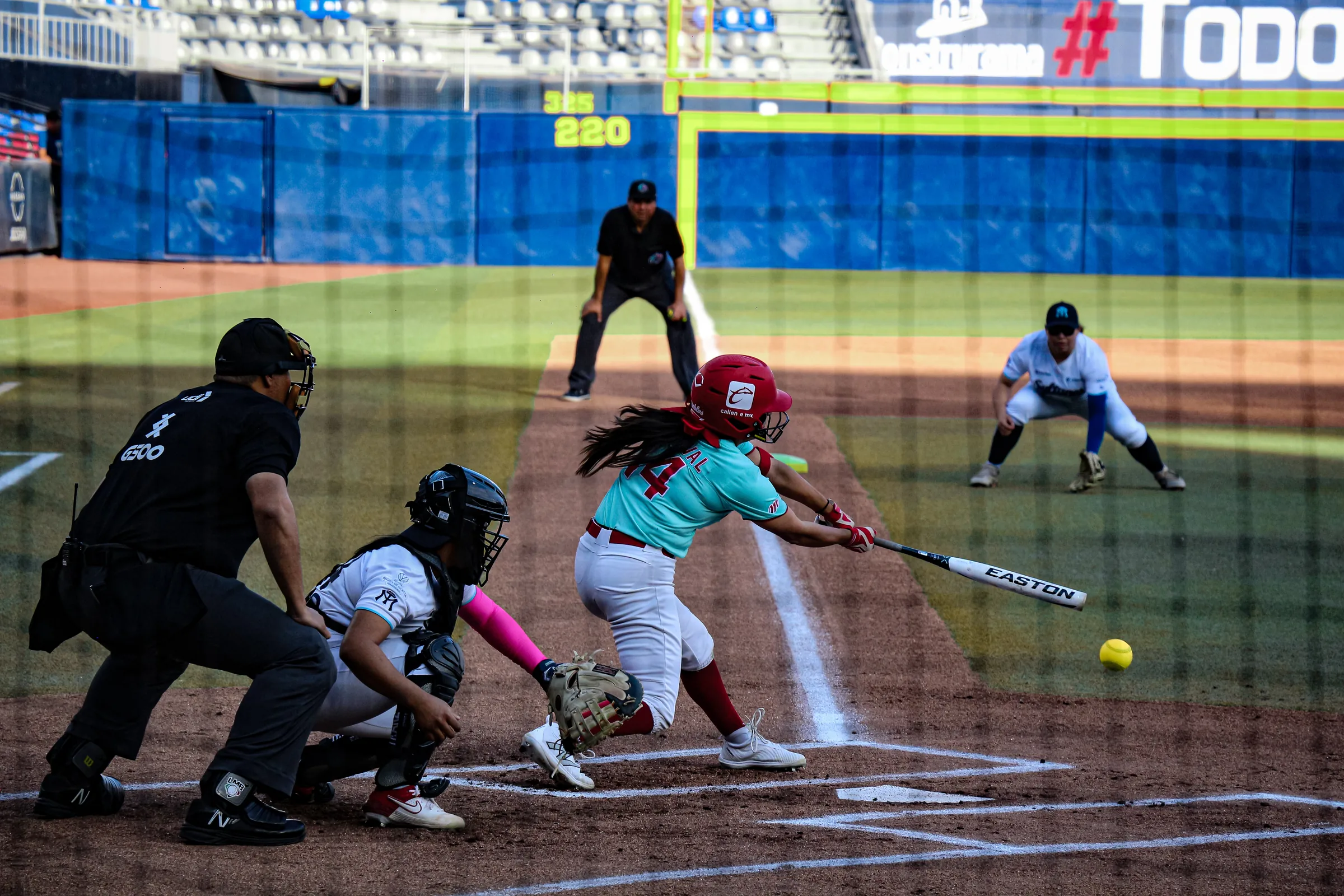 Una bateadora conecta la bola frente al catcher y el ampáyer
