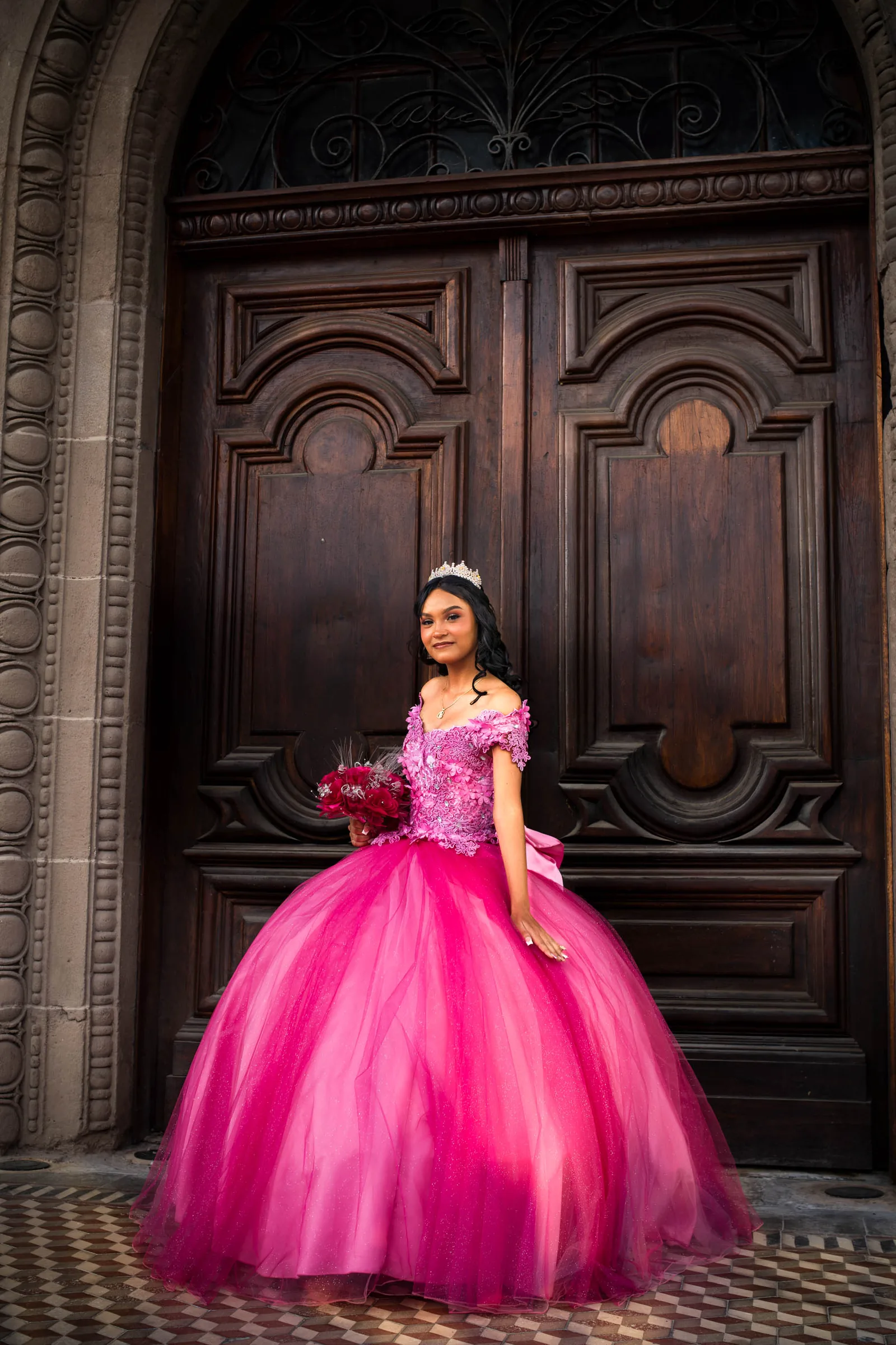 Arleth de pie frente a una puerta de madera tallada con arco de cantera, con el vestido rosa y el ramo de rosas rojas
